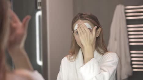 Portrait-of-a-woman-in-bathrobe-putting-white-mask-for-moisturizing-using-fingers