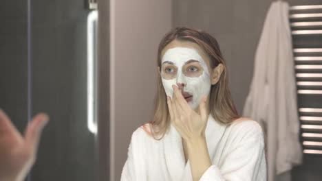 Blonde-woman-in-bathrobe-putting-white-mask-for-moisturizing-using-fingers