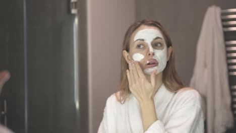 Blonde-woman-in-bathrobe-putting-white-mask-for-moisturizing-using-fingers,-slowmo
