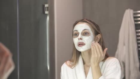 Beautiful-woman-in-bathrobe-putting-white-mask-for-moisturizing-using-fingers,-slowmo