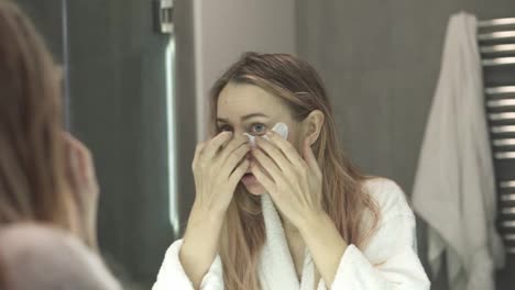 Young-female-in-bathrobe-applying-white-eye-patches-while-looking-at-mirror