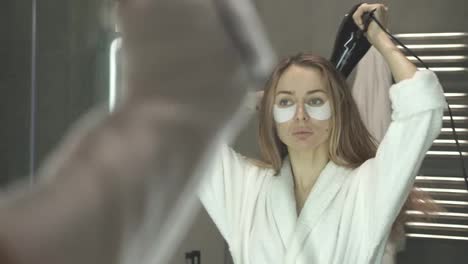 Lady-with-light-hair-and-eye-patches-looking-at-her-reflection-in-bathroom-mirror-and-drying-her-hair-with-hair-dryer