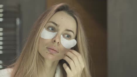 Blonde-woman-in-bathrobe-removing-white-eye-patches-while-looking-at-mirror