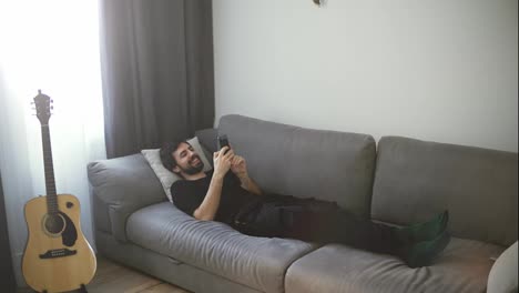 Young-handsome-guy-relaxing-on-sofa-using-mobile,-enjoying-communicating-with-friends