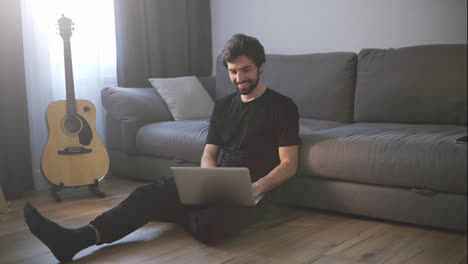 Smiling-man-sitting-on-the-floor-while-video-calling-using-laptop-at-home