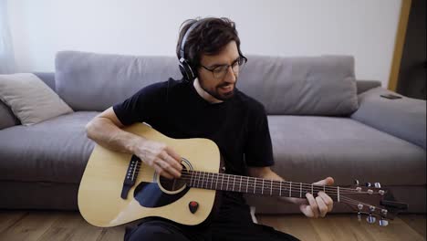 Bearded-man-sitting-on-the-floor,-playing-on-guitar-and-looking-at-the-camera