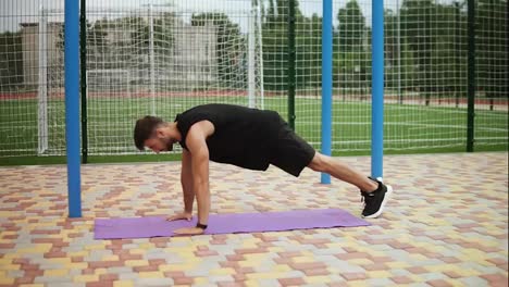 Man-in-black-sportswear-doing-push-ups-on-mat-with-football-field-on-background