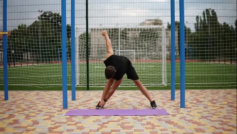 Man-in-black-sportswear-doing-warming-up-exercises-on-mat-with-football-field-on-background,-mill-exercise