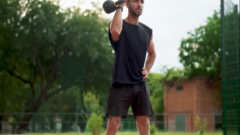 A-bearded-man-in-black-sportswear-trains-with-weight-bob-outdoors-close-to-the-stadium-on-the-racetrack-in-slowmo