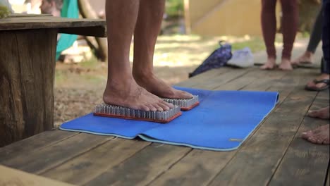 Male-feet-step-on-a-wooden-yoga-board-with-lot-of-metal-nails-pins-around-diverse-unrecognizable-people