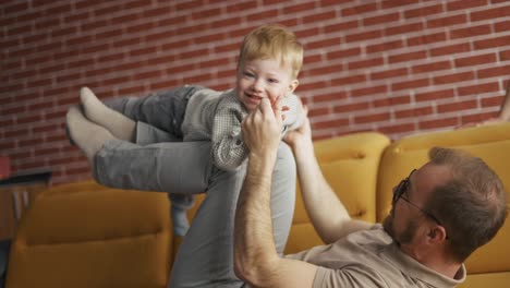 Cheerful-man-holding-jumping-toddler-boy-having-fun-together-on-sofa-in-living-room
