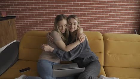Two-happy-friends-with-laptop-sitting-on-a-couch-and-hugging