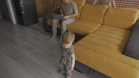 Little-toddler-son-having-fun-at-home-while-their-father-busy-with-laptop-on-the-sofa