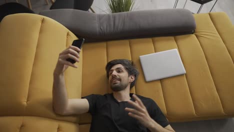 Man-at-home-lying-on-sofa-does-video-call-on-smartphone,-top-view