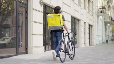Delivery-man-walking-with-bike-and-yellow-bag,-rear-view