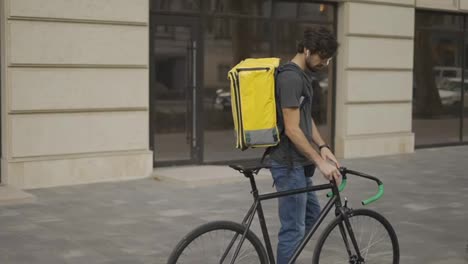 Delivery-man-with-yellow-backpack-walk-on-the-street-with-a-bike,-using-earphones-answer-to-a-call