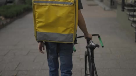 Delivery-man-walking-alone-with-bike-and-yellow-bag,-rear-view