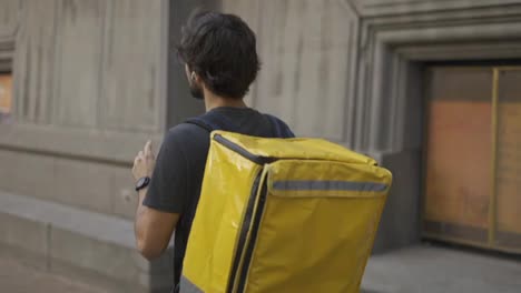 Delivery-man-with-yellow-backpack-talk-to-a-client-on-the-street-using-earphones,-side-view
