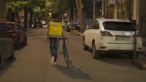 Delivery-man-walking-alone-with-bike-and-yellow-bag-by-evening-city,-rear-view