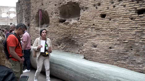 Female-Tour-Guide-Explaining-To-Tourists-About-Marble-Column-Inside-The-Colosseum-In-Rome