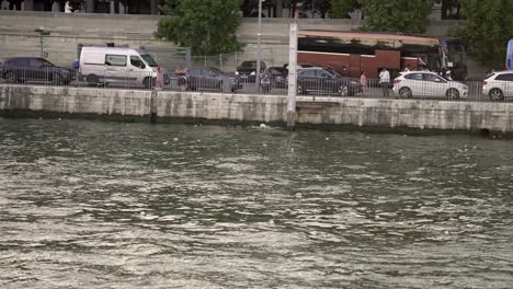 Vehicles-and-people-at-the-shore-of-the-River-Seine,-View-from-boat