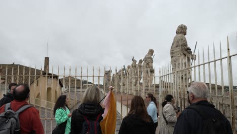 Tourists-On-Rooftop-Of-St-Peter`s-Basilica-Learning-About-The-Statues-Of-Christ-and-Apostles-Seen-Through-Safety-Railings