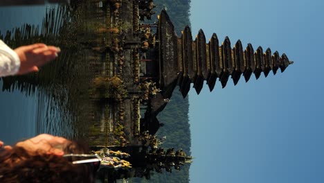 Vertical-ascending-slow-motion-shot-of-a-young-traveler-sitting-on-the-shore-danau-batur-in-front-of-the-water-temple-Pura-Ulun-Danu-temple-overlooking-the-beautiful-lake-with-beautiful-nature