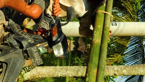 Vertikale-Zeitlupenaufnahme-Eines-Fahrenden-Bauarbeiters-Auf-Einem-Roller-Mit-Bambusstöcken-Auf-Der-Schulter,-Der-Zur-Baustelle-Auf-Bali,-Indonesien-Fährt