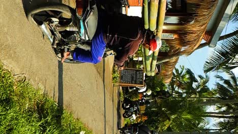 Vertikale-Zeitlupenaufnahme-Eines-Balinesischen-Bauarbeiters,-Der-Mit-Bambusstöcken-Auf-Der-Schulter-Zur-Baustelle-Fährt,-Während-Er-Mit-Seinem-Roller-Fährt