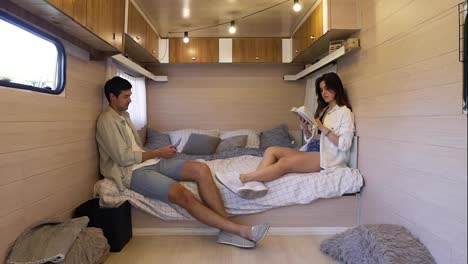 Life-in-the-trailer.-Joint-evening-together-sitting-on-the-bed---attractive,-long-haired-woman-reading-a-book-while-her-husband-scrolling-phone.-Stylish,-modern-interior