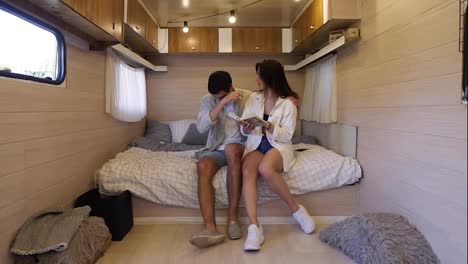 Cheerful,-european-couple-sitting-on-the-big-bed-inside-the-wheels-house,-having-time-together---reading-book,-having-fun.-Recreation,-vacation,-travelling-in-van