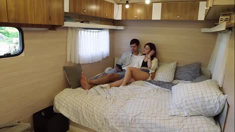 Portrait-of-relaxful-couple-laying-on-the-big-bed-inside-the-wheels-house,-having-time-together---man-reads-book-from-e-reader,-girl-listening-music-with-headphnes.-Recreation,-vacation,-travelling-in-van