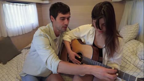 Caucasian,-dark-haired-young-man-teaches-his-girlfriend-to-play-the-guitar-sitting-in-bed-in-wheels-house,-van.-A-romantic-couple-in-love-are-sitting-on-a-bed,-repeating-chords.-Close-up