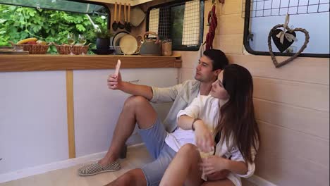 Beautiful-couple-sitting-on-the-floor-in-the-stylish-kitchen-in-van-and-doing-selfie-using-smartphone,-posing-.-Enjoying-togetherness,-shared-holidays,-traveling-by-wheels-house.-Modern-interior