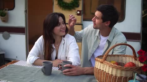 Cheerful-couple-man-and-woman-drinking-coffee-while-sitting-together-at-wooden-table-outdoors.-Man-caressing-hair-of-his-beloved,-bonding.-Stylish,-static-wheels-house-on-background