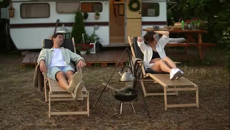 Happy-young-couple-having-rest-close-to-a-campsite-with-hanged-kettle-.-Picnic-of-a-beautiful-couple-in-love-at-the-trailer,-laying-comfortably-on-trestle-beds.-Rest-in-nature.-Modern-trailer-on-background