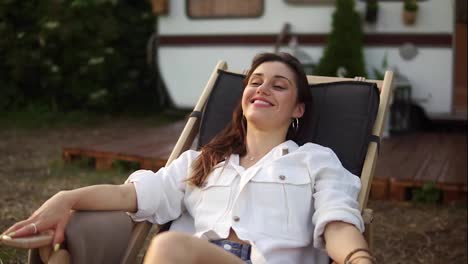 Pleased,-attractive-caucasian-woman-chilling-out-and-smiling-while-laying-on-lounger-outdoors.-Traveling,-rest,-vacation-concept.-Trailer,-wheels-house-on-the-blurred-background