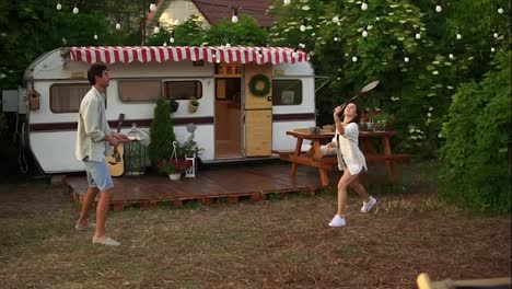 Two-young-tourist-couple-play-badminton-next-to-their-trailer-in-american-retro-style.-Active-vacation,-rest,-traveling.-Summer,-warm-day