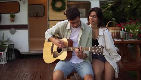 Romantic-couple-sitting-outdoors-in-front-their-home-van-and-man-playing-the-guitar-for-girlfriend.-Outdoor.-Girl-listening-music-and-hugging-man.-Slow-motion