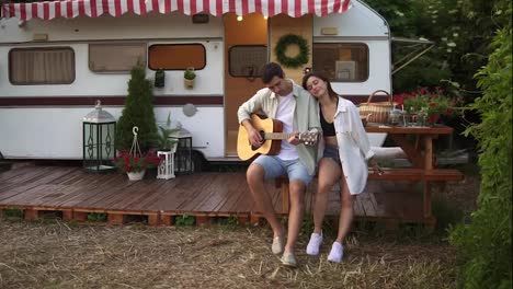 Pleased,-relaxful-couple-sitting-outdoors-in-front-their-home-trailer-and-man-playing-the-guitar-for-girlfriend.-Outdoor.-Girl-listening-music-and-hugging-man.-Slow-motion.-Portrait