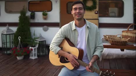 Man-plays-guitar,-holding-acoustic-guitar-and-smiling-to-the-camera.-Sitting-against-the-wheels-house,-trailer-with-nice-exterior-decoration-around.-Portrait-shot