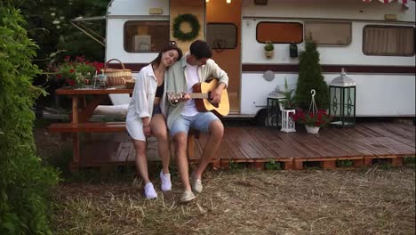 Pleased,-relaxful-couple-sitting-outdoors-in-front-their-home-trailer-and-man-playing-the-guitar-for-girlfriend.-Outdoor.-Girl-listening-music-and-hugging-man.-Slow-motion.-Front-view