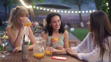Three-elegant-female-friends-sit-in-the-outdoors-cafe,-drink-juice-and-have-fun-communicating