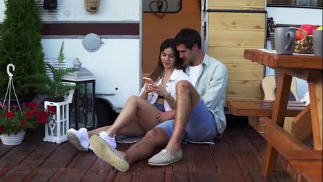 Attractive-couple-sitting-on-the-floor-close-to-the-door-van-and-watching-video-on-smartphone,-smiling-.-Enjoying-togetherness,-shared-holidays,-traveling-by-wheels-house