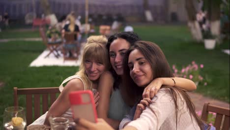 Carefree-women-chatting-in-veranda-cafe-outdoors-taking-selfie-photo-or-video-on-smartphone
