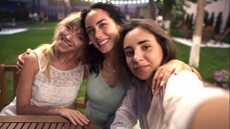 Three-attractive-women-met-in-veranda-cafe-outdoors-taking-selfie-photo-or-video-on-smartphone