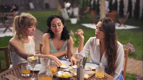 Three-cheerful-women-resting-talking-drinking,-taking-in-summer-veranda-cafe-casual-city-friendship-models-outdoors