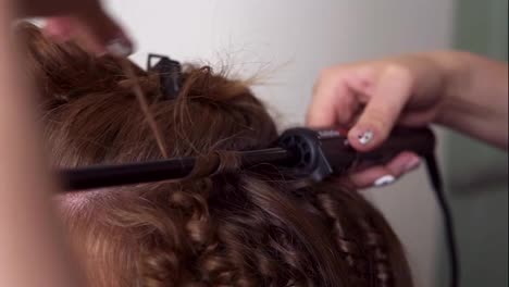 Close-up-shot-of-a-red-headed-woman-getting-her-hairs-curled-in-the-studio.-Hairdressed-uses-curling-iron-stick-applying-hair-on-it.-Unrecognizable