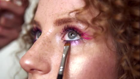 Portrait-of-a-young-woman-during-professional-makeup.-The-makeup-artist-applies-pink-shadows-on-the-girl-s-lower-eye-with-a-brush.-Fashion-and-beauty.-Slow-motion