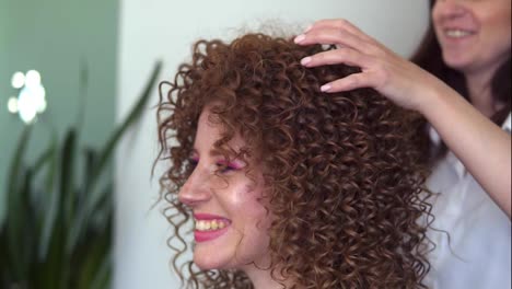 Professional-hair-stylist-finishing-modeling-hairdo-to-young-smilling-girl-customer-with-red-coloured-hair.-Hairdresser-woman-making-final-touch-to-ginger-long-curly-hair